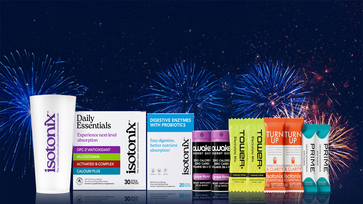 A group of Isotonix dietary supplement products arranged in a row against a dark blue background with fireworks. The products include a white cup, Daily Essentials box, Digestive Enzymes box, two Toucan flavor packets, and two Prime flavor packets.