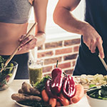 A close-up lifestyle shot of a person preparing a healthy salad, with a plate of fresh vegetables including red bell peppers, celery, and greens, alongside a person chopping ingredients in the background.