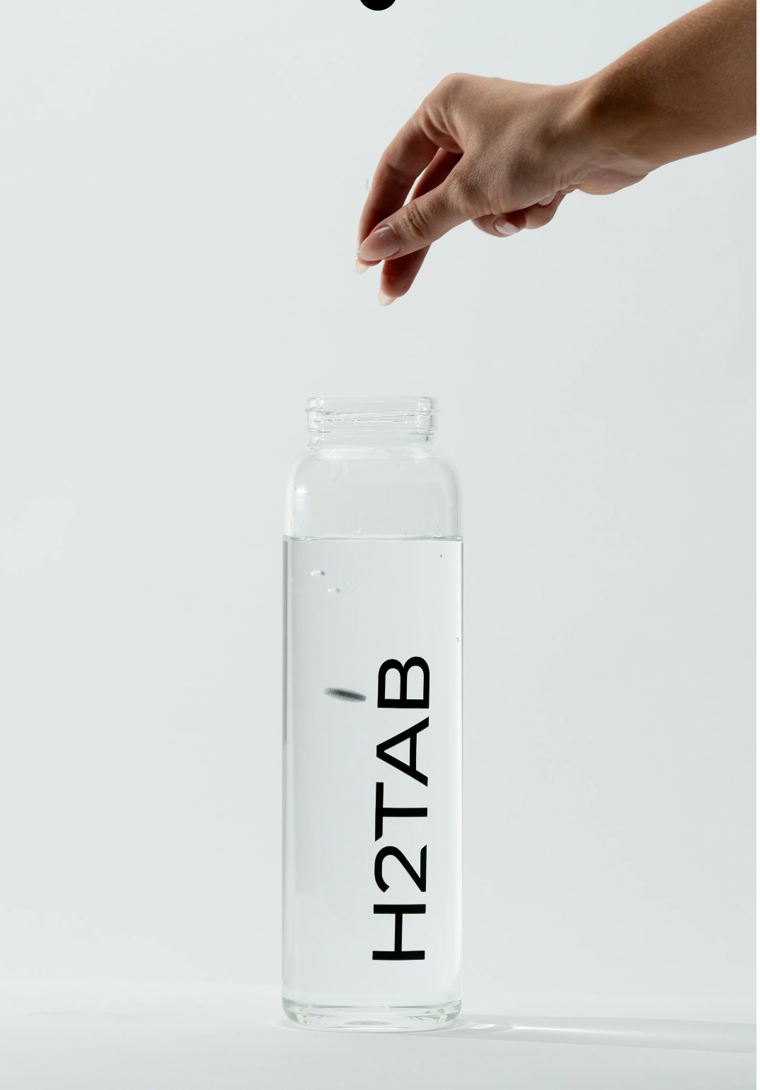 A clear glass water bottle with black vertical text reading H2TAB, shown on a light gray background with a hand reaching toward the opening at the top.