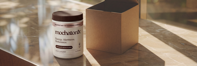 A white jar of mochatonix Energy HarNESS endurance powder with a dark brown lid sits on a polished marble countertop next to an open kraft paper box, with warm sunlight casting shadows in a windowed room.