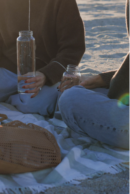 Two people sitting on a blanket on a beach, holding clear glass bottles with a string bubbler setup. A woven straw bag is in the foreground, and the background shows sand and water with warm golden-hour lighting.