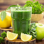 A glass of thick green juice with lemon slices and green herbs and apples around it on a wooden surface.