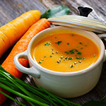 A white ceramic bowl filled with orange carrot soup, garnished with chopped green herbs, set on a rustic wooden surface with whole carrots and green onions around it.