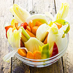 A clear glass bowl filled with a colorful vegetable salad, including celery, carrots, and green onions, set on a rustic wooden surface.