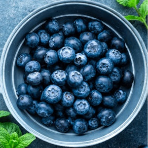 Fresh blueberries in a round metal bowl, photographed from above on a dark textured surface with mint leaves at the corners.