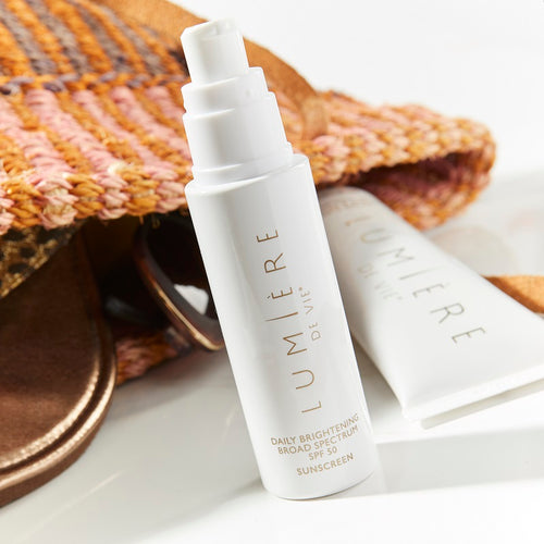 Lumière de Vie Daily Brightening Broad Spectrum SPF 50 pump bottle, next to a straw purse