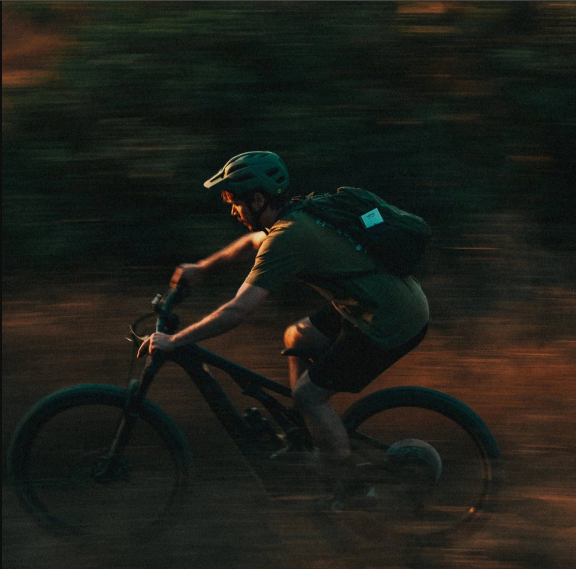 A person riding a bicycle on a dirt path at dusk, wearing a green helmet, olive shirt, dark shorts, and a black backpack, with motion blur in the background.