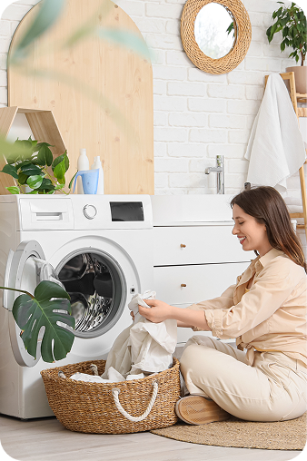 A woman in a beige pajama set sits on a woven rug next to a white front-loading washing machine, folding white clothes from a wicker laundry basket in a bright, modern laundry room.