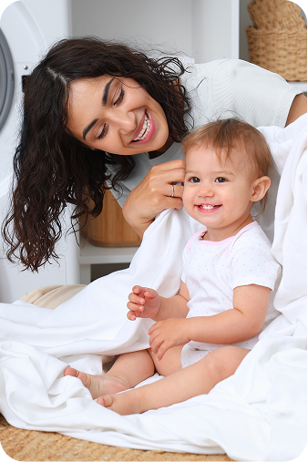 A smiling woman and a baby sitting together on white bedding, both wearing white tops in a bright indoor setting.