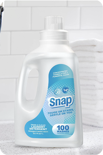 A bottle of Snap Fresh Clean Laundry Detergent, 100 washes, in a white plastic jug with a blue and white label, set against a blurred bathroom tile background.