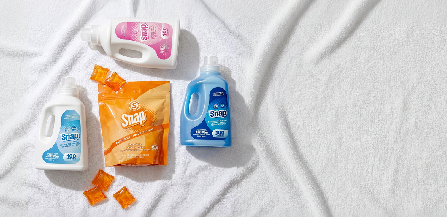 Four Snap laundry care products arranged on a white textured fabric background: a white bottle with pink label, a white bottle with blue label, an orange bag, and two orange sachets.