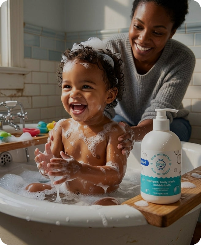 A smiling toddler with soapy hair sits in a bathtub while an adult assists, with a bottle of bubble bath placed on the tub edge in a real bathroom setting.