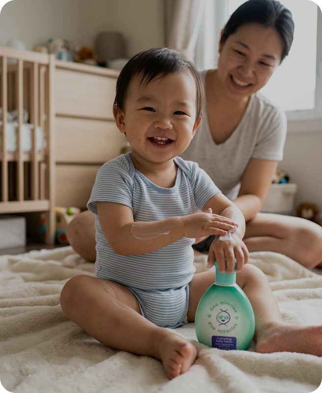 A baby in a blue and white striped onesie sits on a cream blanket in a nursery, smiling while applying lotion from a mint green bottle held by a woman in a white shirt.