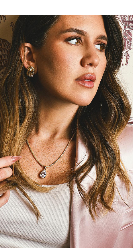 A woman with long wavy brown hair models a delicate gold chain necklace with a small clear teardrop-shaped gemstone pendant. She is wearing a light pink blazer over a ribbed white top, and the background is a soft-focus indoor setting.