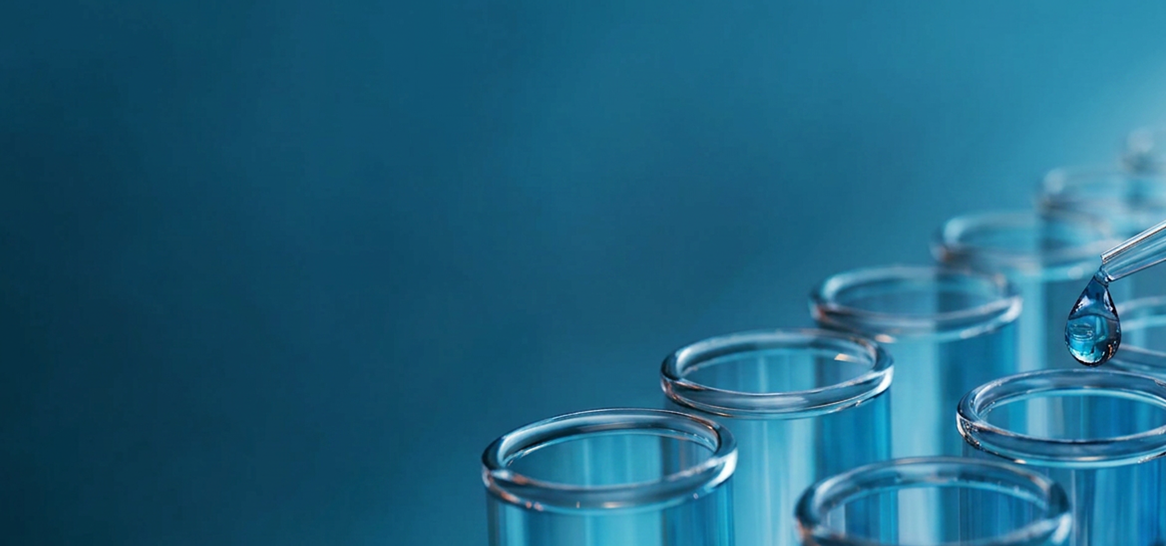 A close-up row of clear glass test tubes on a blue background, with a pipette tip dropping liquid into one of the tubes.