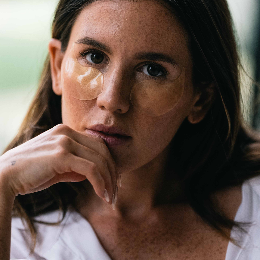 A close-up portrait of a woman with two clear under-eye patch masks applied to her cheeks, resting her chin in her hand. She has freckles and is wearing a white top, set against a soft, blurred background.