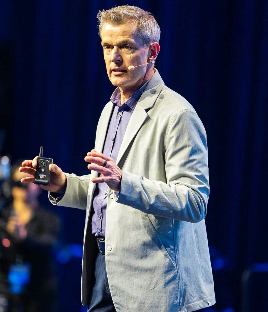 A man giving a presentation on stage, wearing a light gray blazer over a purple button-down shirt, holding a small wireless microphone transmitter against a dark blue curtain background.