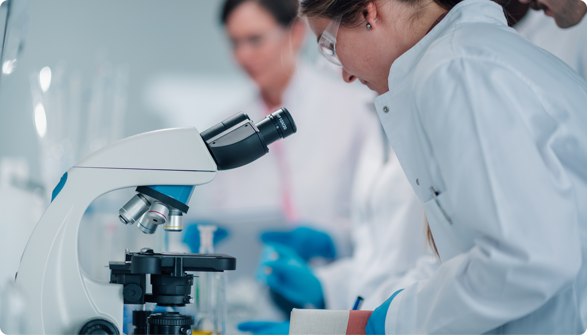 A close-up of a scientist in a white lab coat looking through a microscope in a bright laboratory setting, with another person blurred in the background.