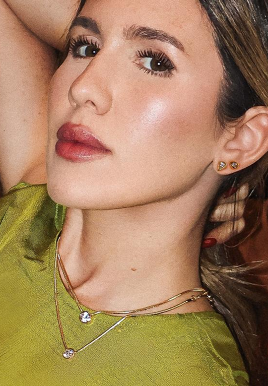 Close-up of a woman wearing delicate layered necklaces and small gold stud earrings, with warm lighting and a soft-focus background.