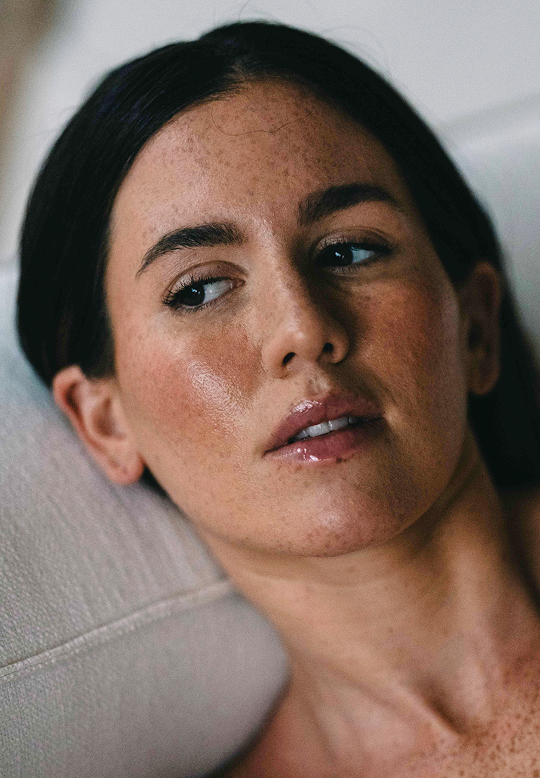 A close-up portrait of a woman with dark hair resting her head on a light beige pillow, looking off-camera with a neutral expression.