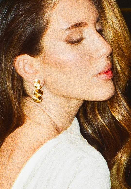 A close-up side profile of a woman with closed eyes wearing a gold spiral drop earring, with her brown hair and white top visible against a dark background.