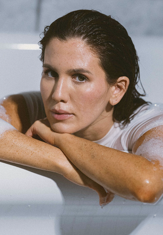 A close-up lifestyle shot of a woman with wet, dark hair resting her head on her arms in a white bathtub filled with soapy water. She is wearing a white t-shirt.