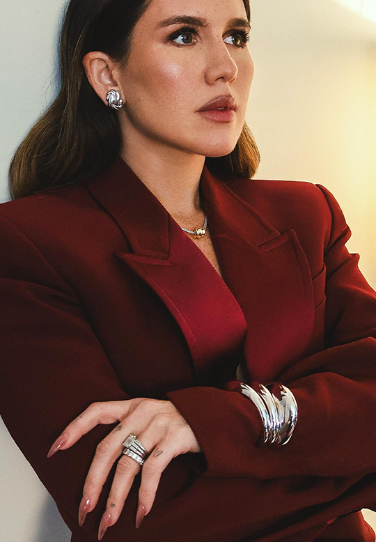 A woman wearing a deep burgundy blazer and matching top, accessorized with silver rings and bracelets, photographed against a soft beige background.