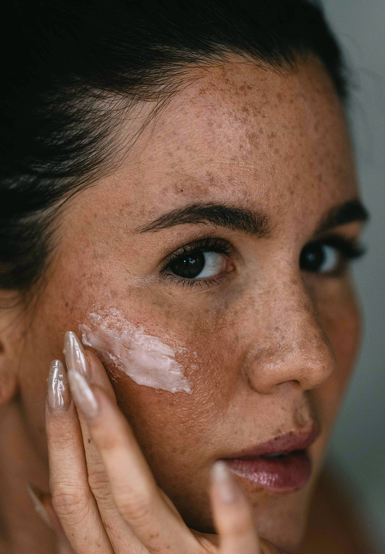 Close-up of a woman applying a white cream or balm to her cheek with her hand, showing her skin texture and freckles against a soft gray background.