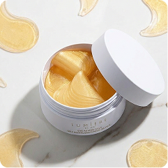 A white cosmetic jar filled with yellow sheet-mask patches, shown open with the lid leaning against it on a light marble surface with scattered mask slices.
