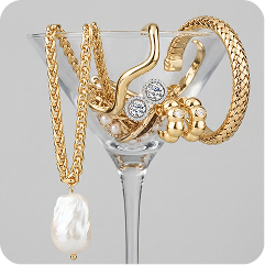 A gold-toned jewelry set draped over a clear martini glass, including a chain necklace with a white pearl drop, bracelets, rings, and a clutch-style bag with clear stones, on a light gray background.