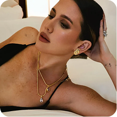 A woman wearing layered gold necklaces and matching gold earrings, lying on a white sofa. She is wearing a black bralette, and the jewelry is the apparent product being showcased.