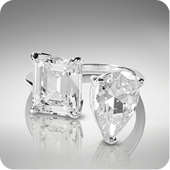 A pair of silver-toned rings featuring a square emerald-cut clear gemstone and a pear-cut clear gemstone, displayed on a reflective gray surface against a gradient gray background.