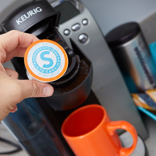 Shopping Annuity Brand Premium Coffee pod shown over coffee maker and an orange cup
