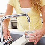 A woman in a yellow top fills a clear glass with running water from a chrome faucet in a kitchen setting.