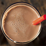 A close-up of a frothy light brown beverage in a clear glass with a red straw, photographed from above against a dark background.