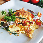 A close-up of a plated breakfast dish featuring golden scrambled eggs mixed with tomato sauce, served with greens and a side of bacon on a white plate.