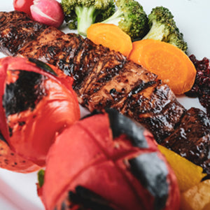 Grilled chicken breast with charred tomatoes, broccoli, and carrots on a white plate.