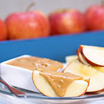 Slices of apple topped with peanut butter in a small white dish, with whole apples blurred in the background on a blue surface.