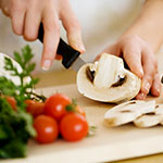 A close-up lifestyle shot of a person slicing a white mushroom with a black-handled knife on a light wooden cutting board, with fresh tomatoes and parsley in the foreground.