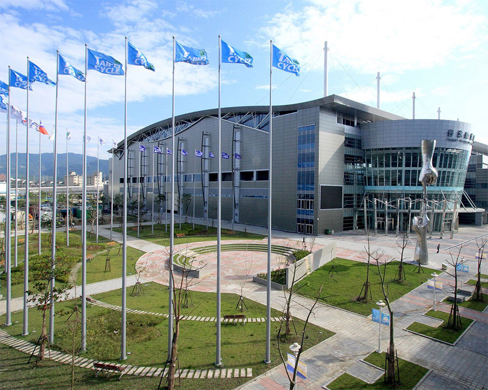 A large modern gray exhibition hall or convention center with glass facade, surrounded by many flagpoles flying blue flags, landscaped green lawn, paved walkways, and a partly cloudy sky.