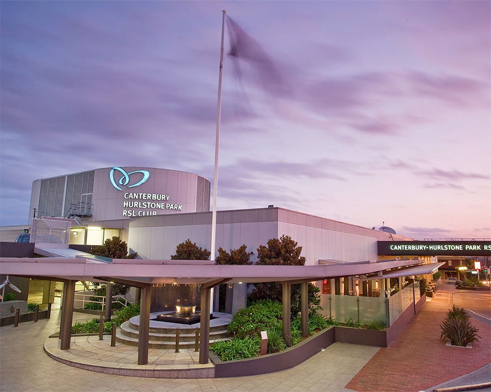 Canterbury-Mount Stone Park & Club building exterior at dusk, with a flagpole, landscaped walkway, covered entrance, and purple-pink sky.