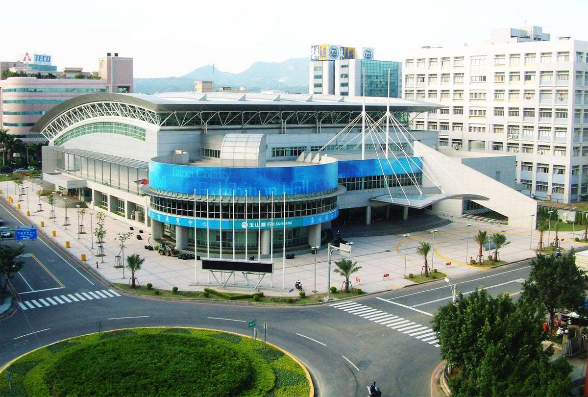 An aerial view of a modern hospital or research building with a curved blue glass facade, surrounded by roads, landscaping, and other buildings in the background.