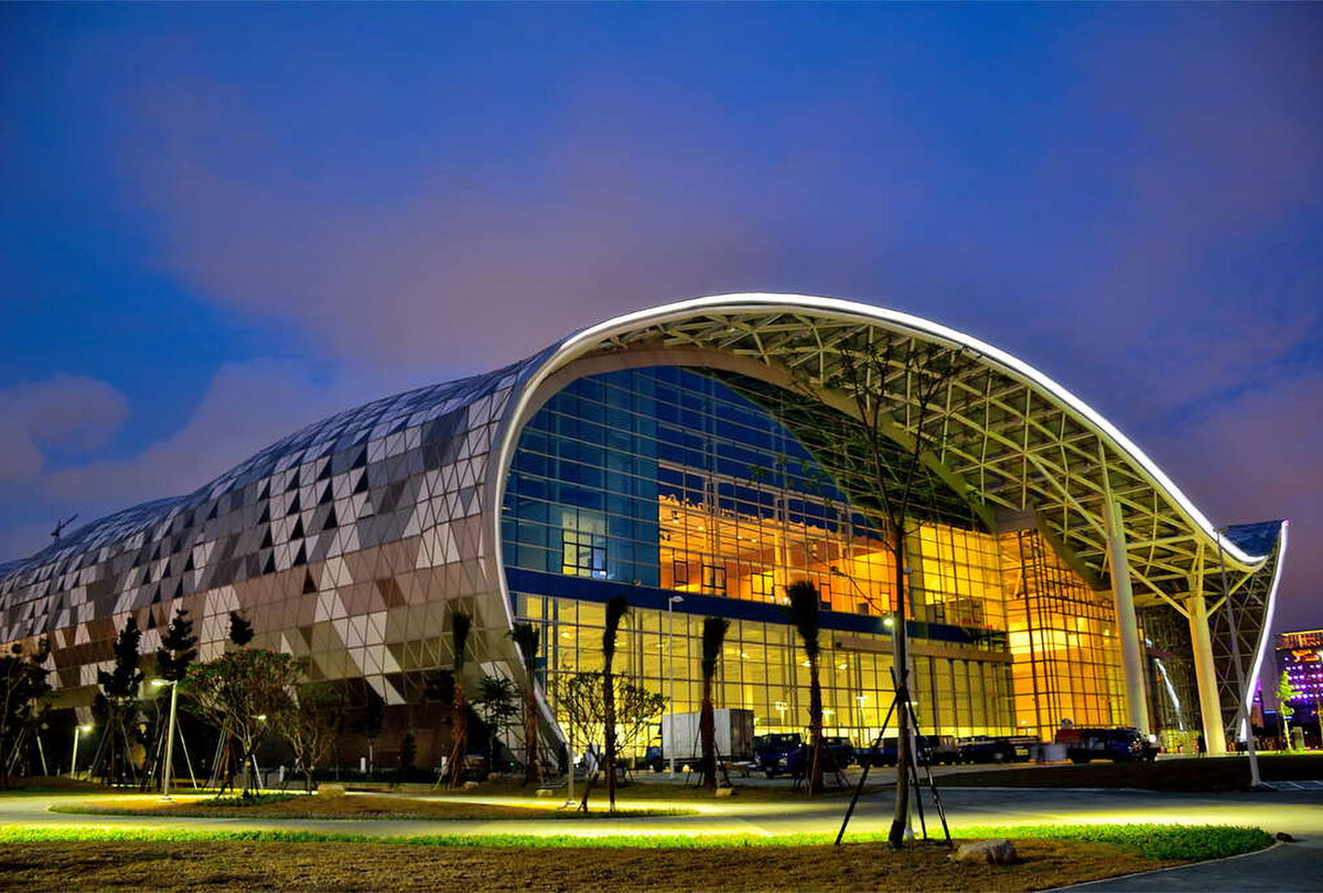 Modern architectural exhibition hall with a curved glass facade and illuminated warm interior, photographed at dusk against a blue sky.