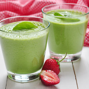 Two clear glasses filled with a bright green smoothie, garnished with mint leaves, set on a white wooden surface with fresh strawberries and a red-and-white checkered cloth in the background.