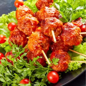 Succulent meatballs glazed in a rich red tomato sauce, served on skewers over a bed of green lettuce and cherry tomatoes on a black plate.