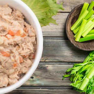 A white bowl filled with shredded canned tuna salad, shown on a rustic wooden table with celery sticks in a small wooden dish and fresh green lettuce leaves.