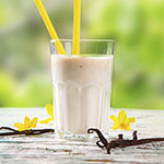 A clear glass filled with a pale yellow creamy beverage, with two yellow straws, set on a rustic wooden surface with small yellow flowers and vanilla beans, against a blurred green outdoor background.