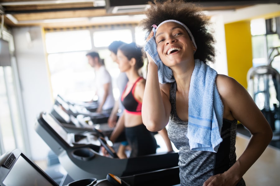 A woman in a gym wearing a gray sports tank top, black leggings, a white headband, and a light blue towel over her head, smiling and wiping sweat. Other gym-goers are visible on treadmills in the background.