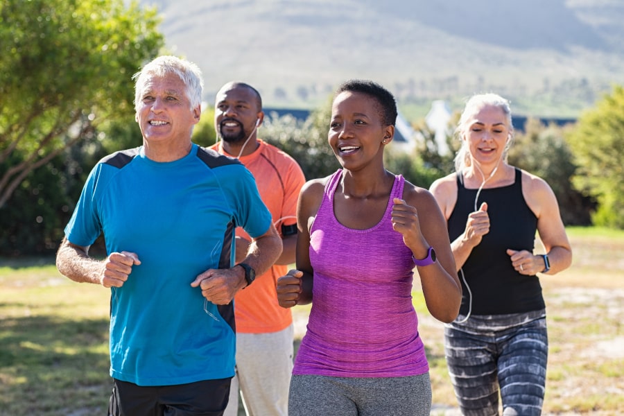 Four smiling fitness enthusiasts jogging together outdoors on a sunny day, wearing athletic wear in a grassy park with trees and hills in the background.