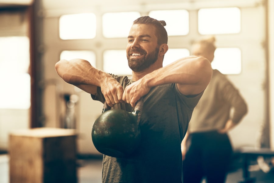 A smiling man in a gym holding a black kettlebell, wearing a dark gray t-shirt. Another person is blurred in the background near windows.